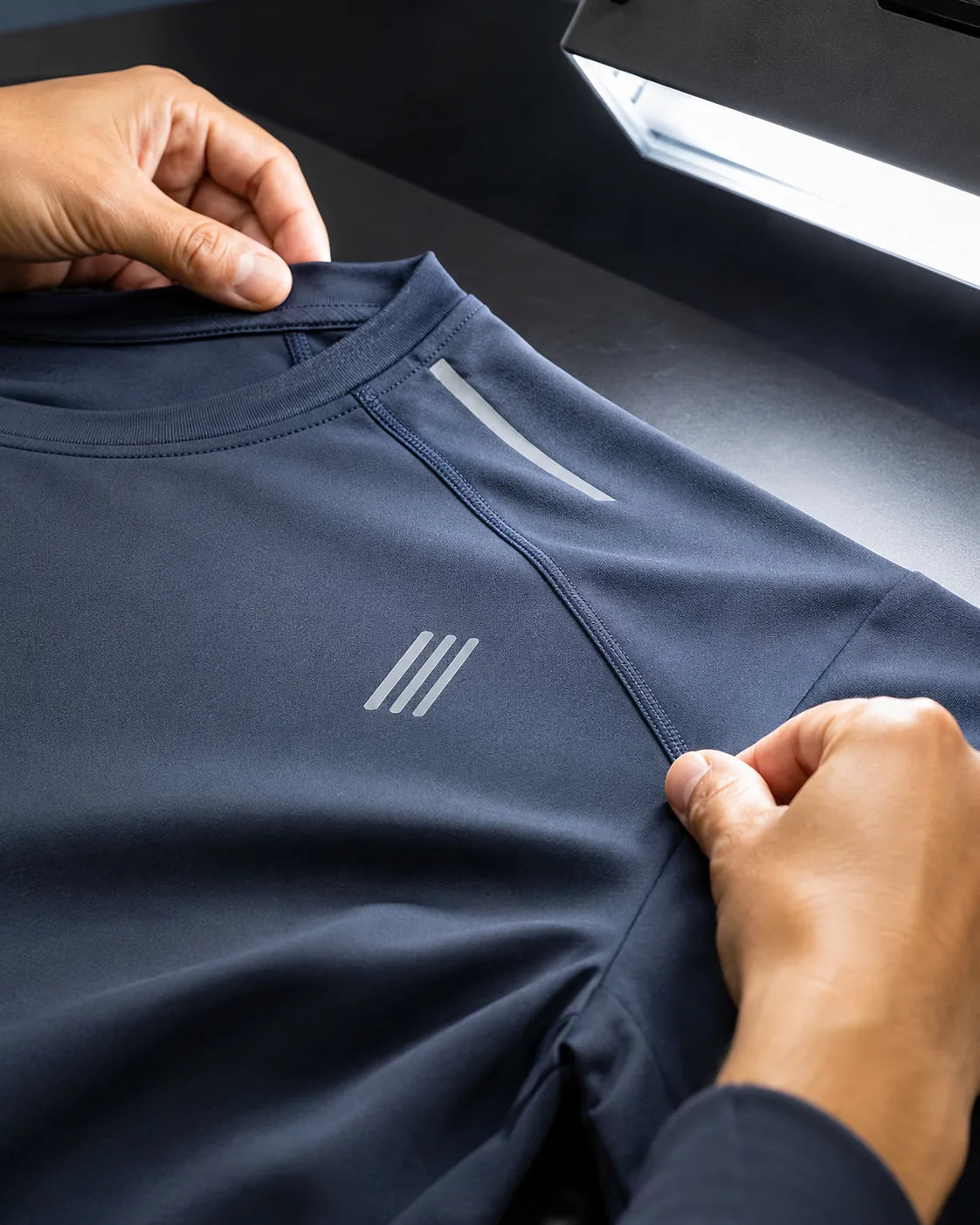 Two hands stretch the shoulder seam of a navy athletic shirt under an inspection lamp