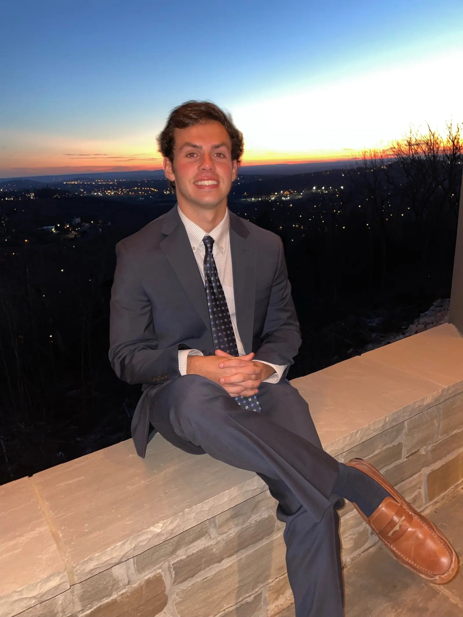 Charlie Reeves in a suit, seated on a stone wall at dusk