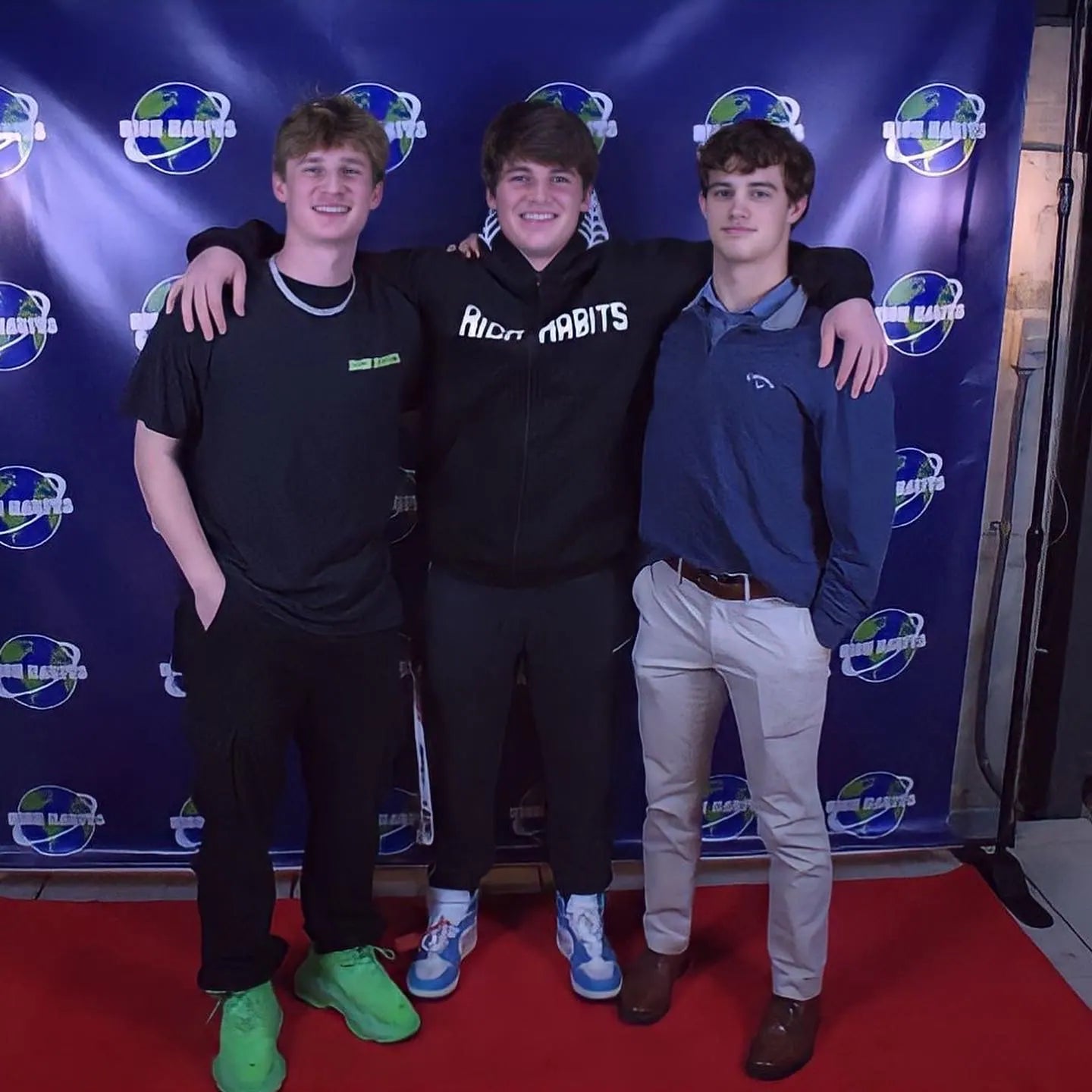 Carter Vail, Sam Sutton and Sam Carr with their arms around each other on a red carpet in front of the original Rich Habits globe step-and-repeat