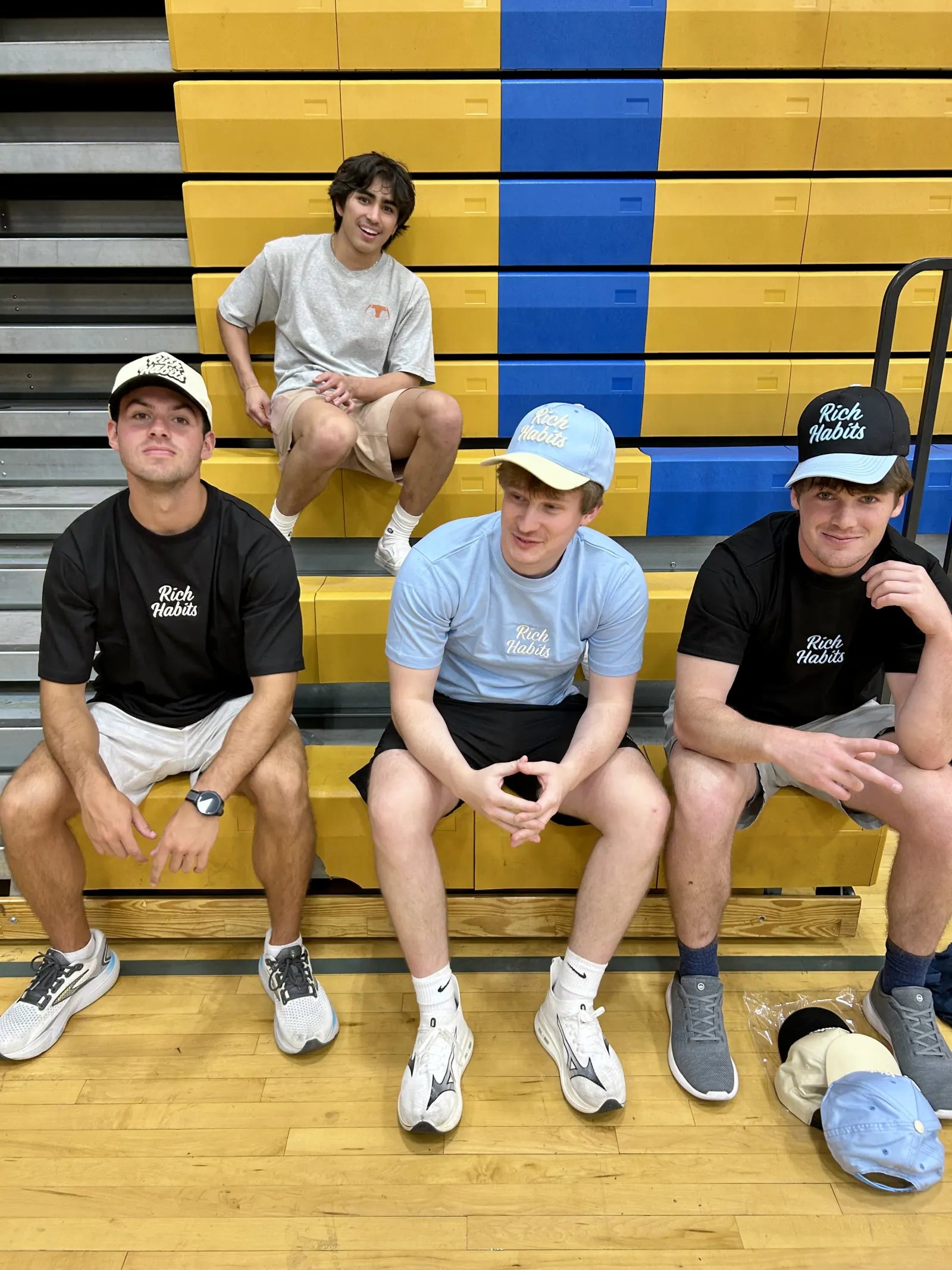 Carter Vail, Sam Carr, Charlie Reeves and Nico Egbalic on gym bleachers in Rich Habits tees and caps