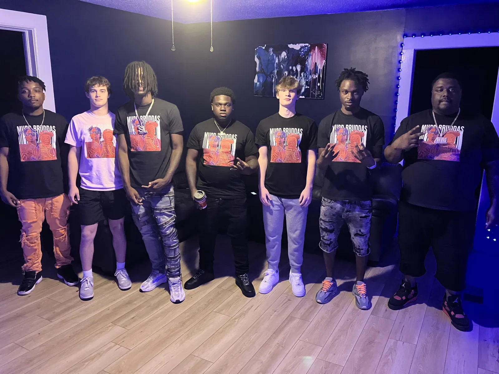 Seven people in Mudd Broddas tees under blue light on a music-video set, Sam Sutton and Carter Vail among them