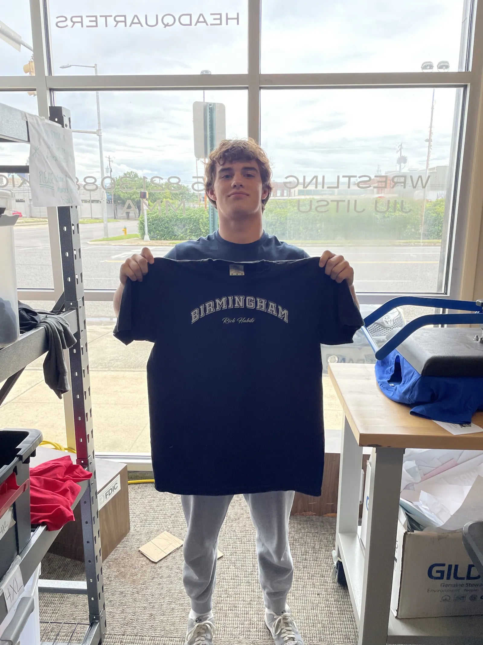 Sam Sutton holding the navy Birmingham tee up in a window
